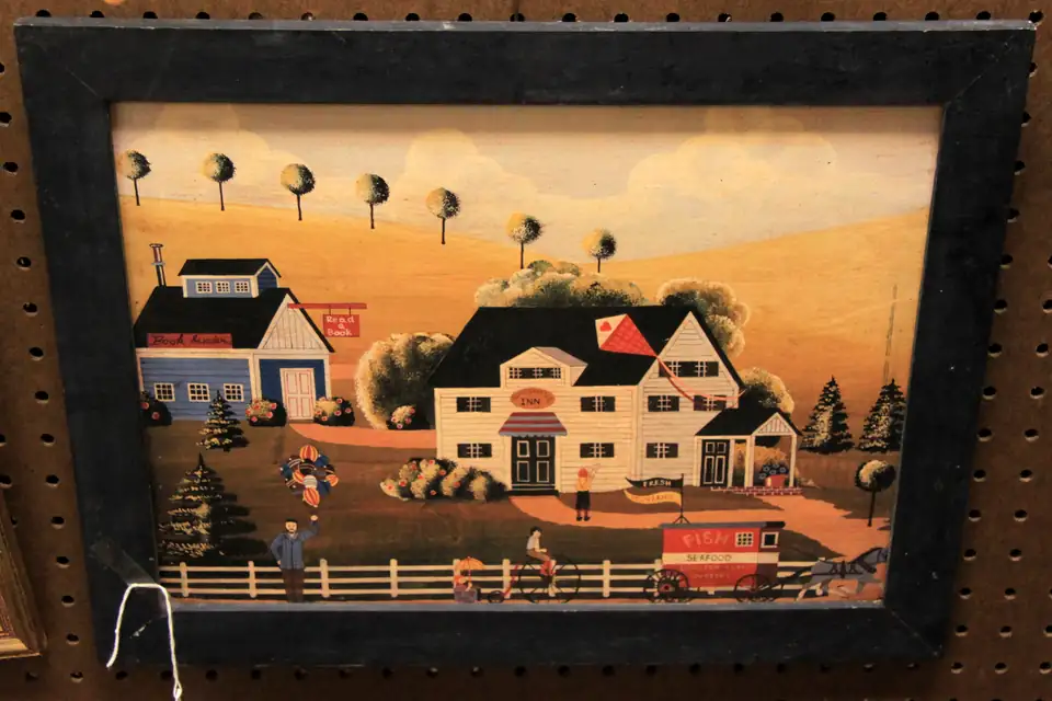 A framed folk-art farm painting hanging on pegboard in an antiques booth.