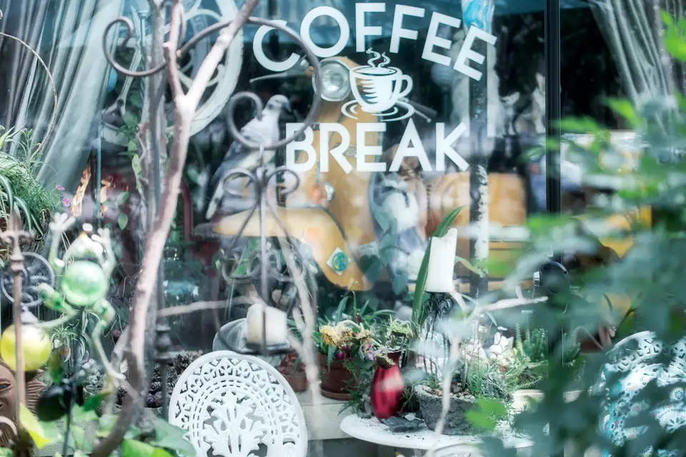 A shop window lettered 'Coffee Break' filled with garden decor, candles and a vintage yellow scooter.
