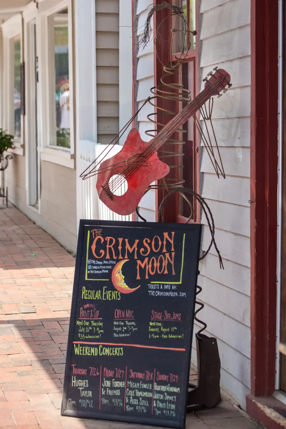 A chalkboard sign for The Crimson Moon and a red guitar sculpture on a brick sidewalk.