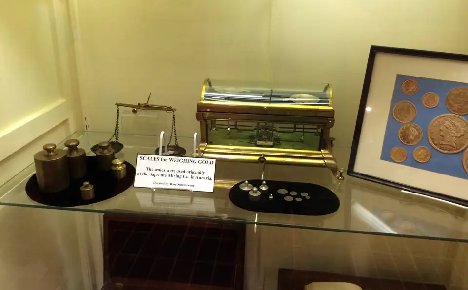 Antique brass scales for weighing gold in a museum display case beside framed gold coins.