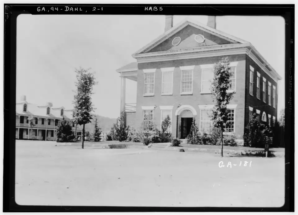 A 1930s black-and-white photograph of the brick Lumpkin County Courthouse on a bare dirt square, with the old hotel behind it.