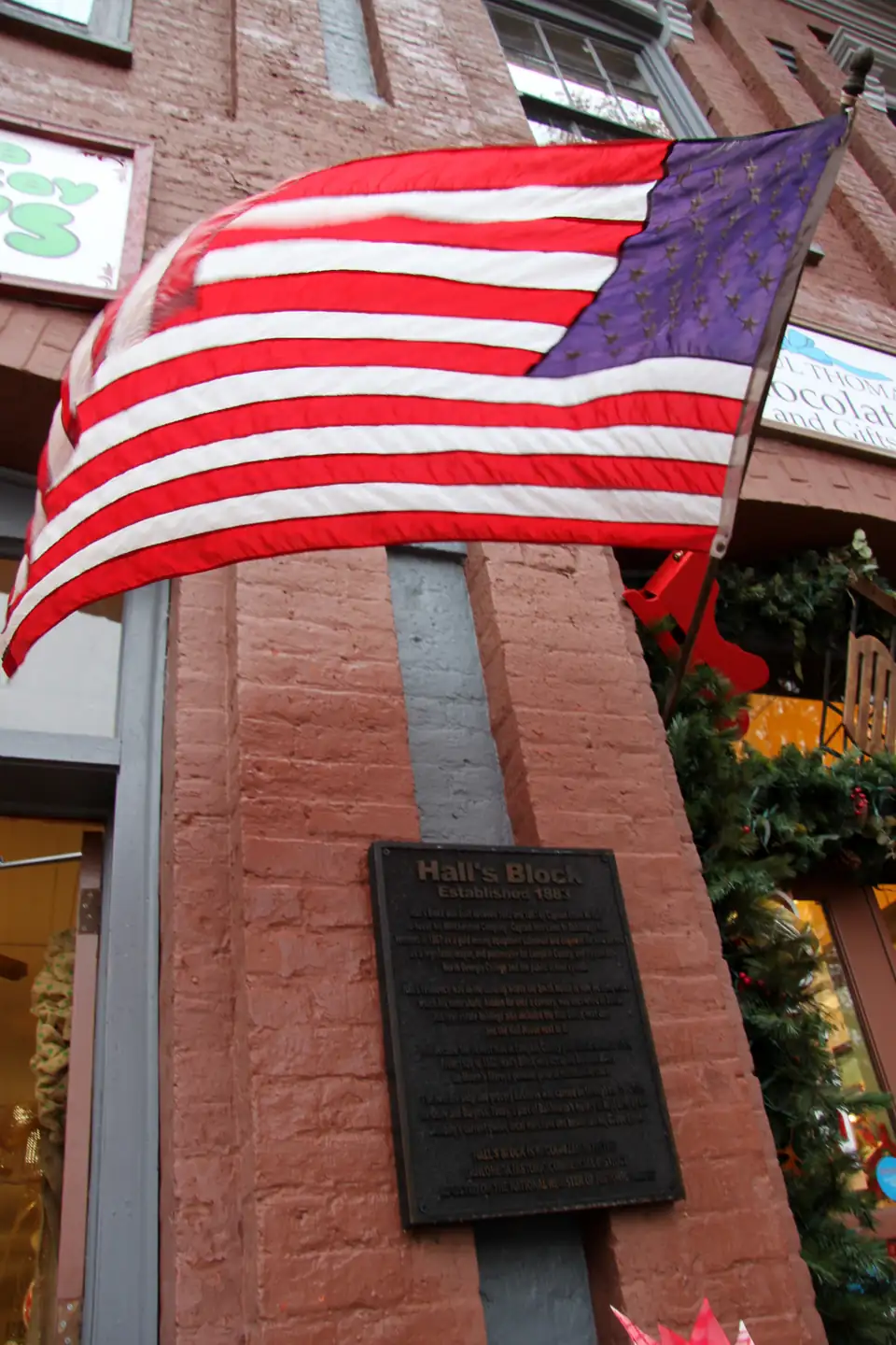 An American flag flying beside the bronze Hall's Block plaque on a brick storefront, with holiday greenery in the window.
